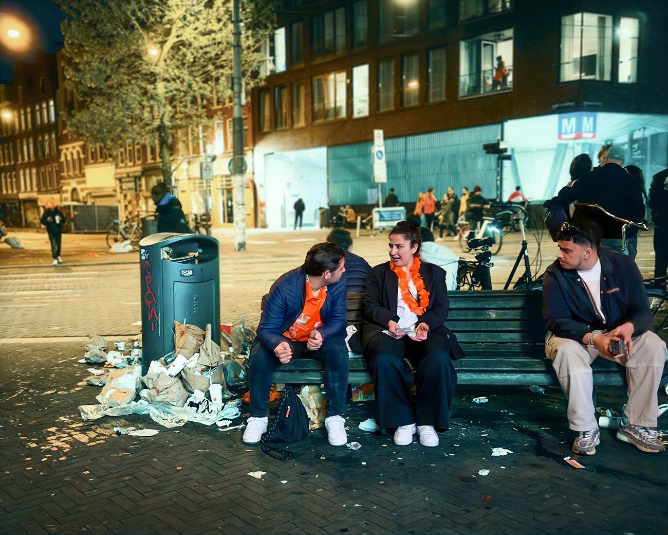 Straatbeeld in Amsterdam na Koningsdag, met mensen op een bankje, omringd door afval bij een overvolle prullenbak, en feestelijke oranje kleding.