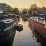 Boten in een rustige Amsterdamse gracht bij zonsondergang, met moderne en traditionele gebouwen op de achtergrond.