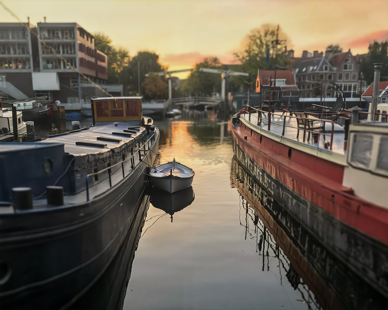 Boten in een rustige Amsterdamse gracht bij zonsondergang, met moderne en traditionele gebouwen op de achtergrond.