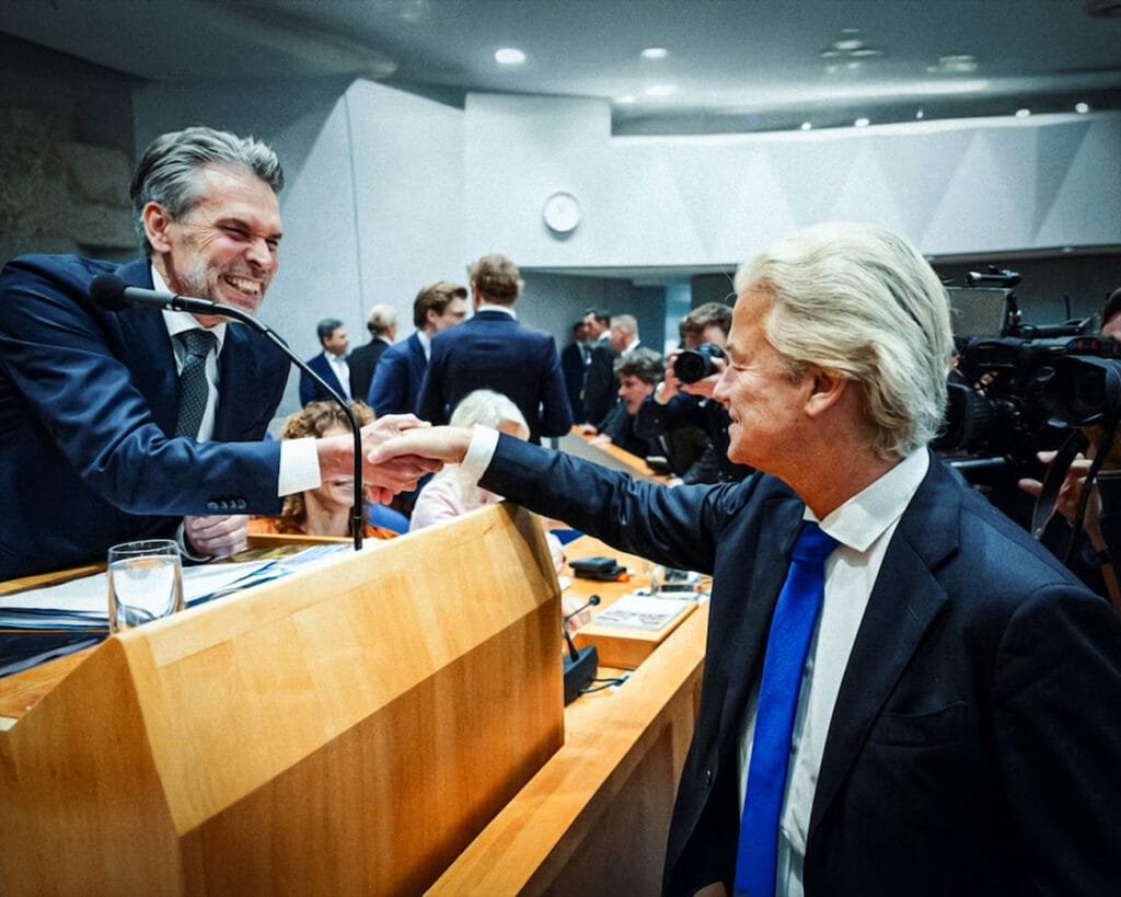 Minister Schoof schudt de hand van Geert Wilders tijdens een debat in de Tweede Kamer, een symbolisch moment dat de onderlinge samenwerking en normalisering van extreemrechts in Nederland illustreert.