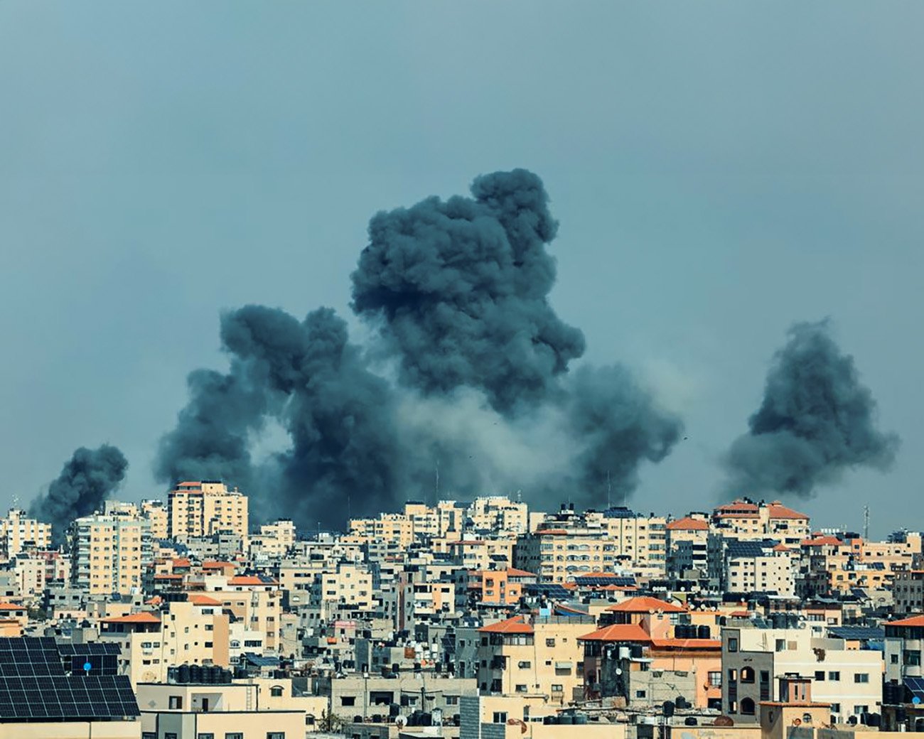 Rookpluimen stijgen op boven Gaza-stad na bombardementen, zichtbaar tegen een sombere lucht met verwoeste gebouwen op de achtergrond.