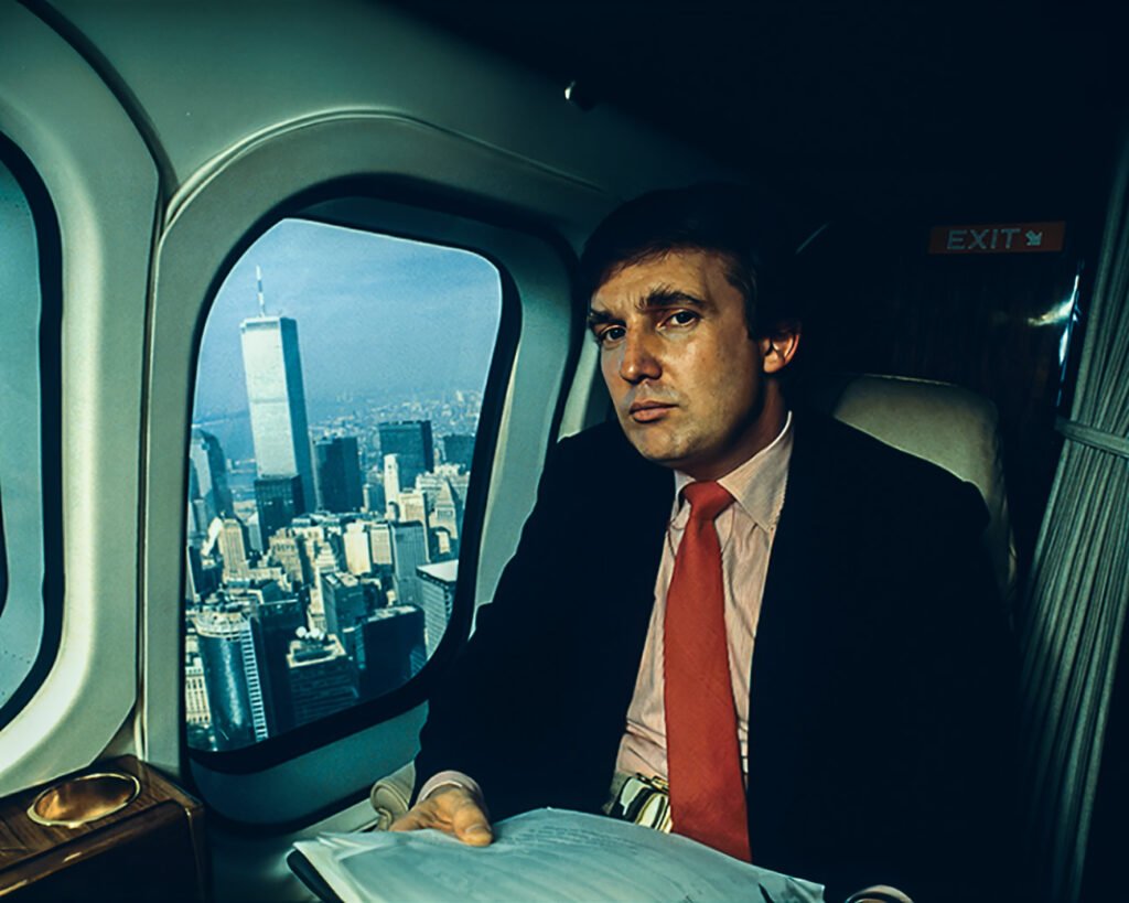 Donald Trump in de jaren ’80, zittend in zijn privéjet terwijl hij over Manhattan vliegt. De skyline van New York is zichtbaar door de ramen, wat een beeld schetst van zijn rijke levensstijl en ambities in die tijd.