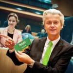 Geert Wilders toont een groene sticker met anti-islam boodschap tijdens een parlementaire vergadering, geflankeerd door collega’s.