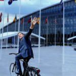 Mark Rutte op een fiets voor het NAVO-hoofdkwartier, zwaaiend naar de camera.