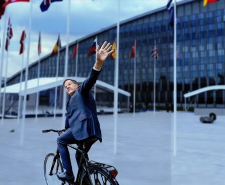 Mark Rutte op een fiets voor het NAVO-hoofdkwartier, zwaaiend naar de camera.