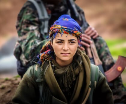 Portret van een jonge vrouwelijke strijder uit Rojava, gekleed in militaire kleding met een kleurrijke hoofddoek, symbool van verzet en solidariteit.