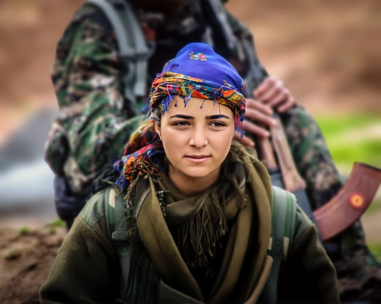 Portret van een jonge vrouwelijke strijder uit Rojava, gekleed in militaire kleding met een kleurrijke hoofddoek, symbool van verzet en solidariteit.