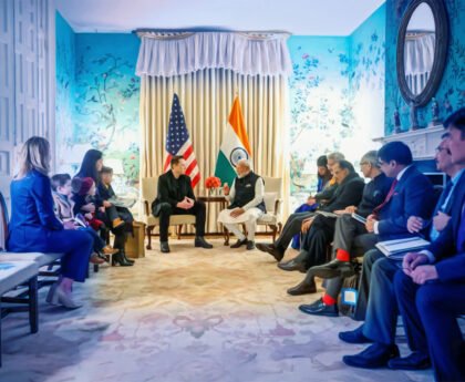 Elon Musk en Narendra Modi in diplomatieke ontmoeting, omringd door kinderen van Musk en Indiase delegatie, in een formele setting met Amerikaanse en Indiase vlag.