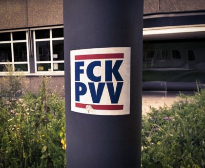 Sticker met de tekst “FCK PVV” op een lantaarnpaal, met op de achtergrond een gebouw en groenstrook.