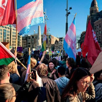 Een demonstratie op de Dam in Amsterdam met rode socialistische vlaggen en transgender vlaggen, waarbij mensen protesteren voor sociale rechtvaardigheid en transrechten.