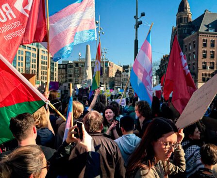 Een demonstratie op de Dam in Amsterdam met rode socialistische vlaggen en transgender vlaggen, waarbij mensen protesteren voor sociale rechtvaardigheid en transrechten.