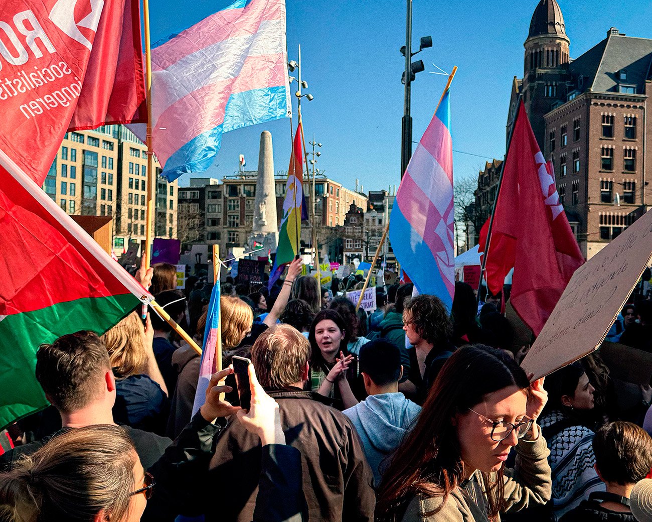 Een demonstratie op de Dam in Amsterdam met rode socialistische vlaggen en transgender vlaggen, waarbij mensen protesteren voor sociale rechtvaardigheid en transrechten.