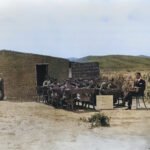 Historische foto van een openluchtklas bij een eenvoudige school van klei, met kinderen aan houten bankjes en een leraar in pak. Een krijtbord toont een boodschap over onderwijs, gedateerd 1920. De school bevindt zich in een landelijke omgeving.