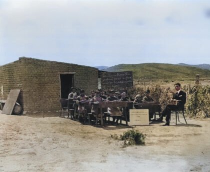 Historische foto van een openluchtklas bij een eenvoudige school van klei, met kinderen aan houten bankjes en een leraar in pak. Een krijtbord toont een boodschap over onderwijs, gedateerd 1920. De school bevindt zich in een landelijke omgeving.