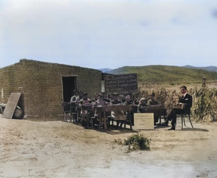 Historische foto van een openluchtklas bij een eenvoudige school van klei, met kinderen aan houten bankjes en een leraar in pak. Een krijtbord toont een boodschap over onderwijs, gedateerd 1920. De school bevindt zich in een landelijke omgeving.