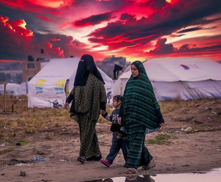 Twee Palestijnse vrouwen in traditionele kleding lopen met een kind door een vluchtelingenkamp bij zonsondergang, met dramatische rode luchten op de achtergrond.