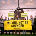 Een groep Greenpeace-activisten protesteert met een spandoek waarop staat “We Will Not Be Silenced”, met op de achtergrond een Nederlandse windmolen.