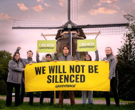 Een groep Greenpeace-activisten protesteert met een spandoek waarop staat “We Will Not Be Silenced”, met op de achtergrond een Nederlandse windmolen.