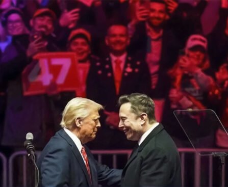 Trump en Musk schudden elkaar de hand tijdens een campagnebijeenkomst, omringd door juichende aanhangers met een bord met nummer 47.