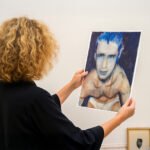 Persoon bekijkt een portret van een halfnaakte, kwetsbare man met blauwe haren in een kunstgalerie.