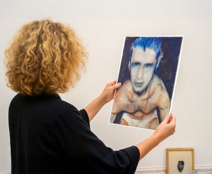 Persoon bekijkt een portret van een halfnaakte, kwetsbare man met blauwe haren in een kunstgalerie.