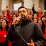 Zohran Mamdani spreekt een menigte toe tijdens een demonstratie, omringd door supporters in rode kleding