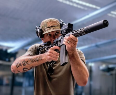 Pete Hegseth richt een semi-automatisch geweer terwijl zijn “We the People”-tatoeage zichtbaar is op zijn onderarm.