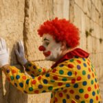 Clown in kleurrijk kostuum die met zijn handen tegen de Klaagmuur leunt.