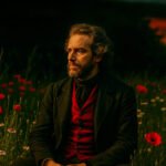 Portret van een man in donkere kleding zittend in een veld met klaprozen bij zonsondergang.