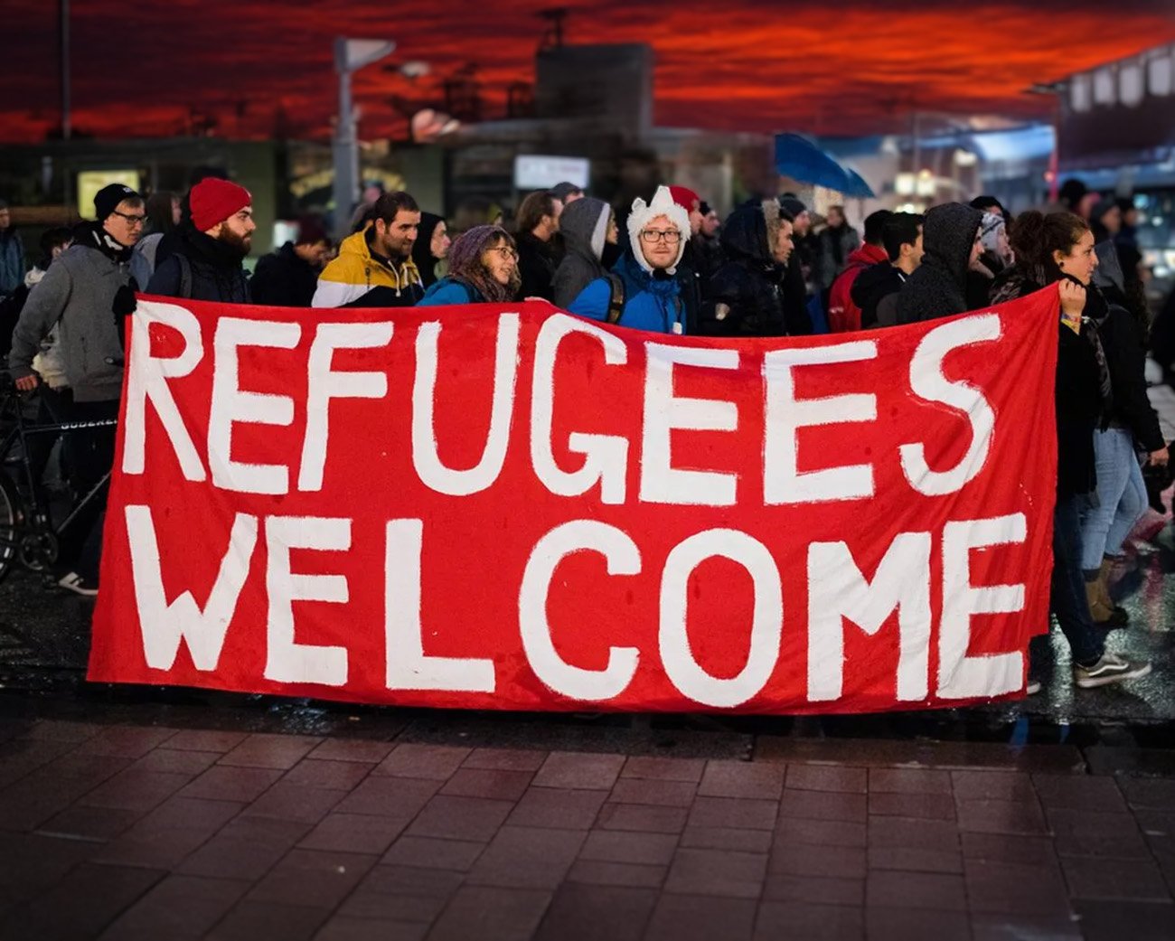 Groep mensen met spandoek ‘Refugees Welcome’ tijdens demonstratie bij zonsondergang.