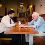 Zohran Mamdani en Bernie Sanders zitten tegenover elkaar aan een tafel in een restaurant in Astoria en voeren een betrokken gesprek.