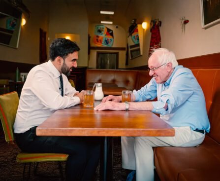 Zohran Mamdani en Bernie Sanders zitten tegenover elkaar aan een tafel in een restaurant in Astoria en voeren een betrokken gesprek.