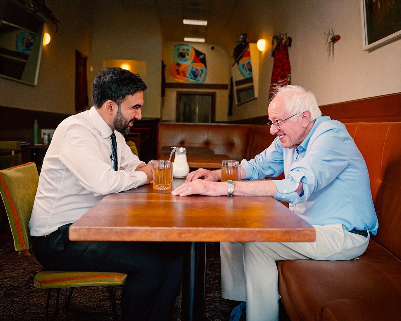 Zohran Mamdani en Bernie Sanders zitten tegenover elkaar aan een tafel in een restaurant in Astoria en voeren een betrokken gesprek.