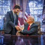 Mamdani en Trump schudden elkaar de hand in het Oval Office, één achter het bureau en de ander staand ernaast, omringd door vlaggen en foto’s.