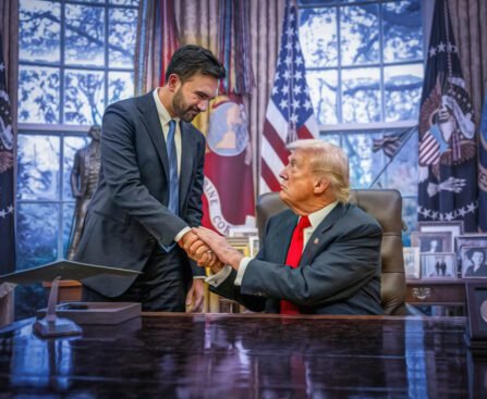 Mamdani en Trump schudden elkaar de hand in het Oval Office, één achter het bureau en de ander staand ernaast, omringd door vlaggen en foto’s.