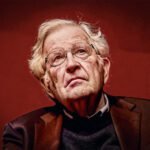 Noam Chomsky kijkt iets omhoog met een rustige, nadenkende blik, zittend tegen een effen rode achtergrond, met bril en grijs haar.