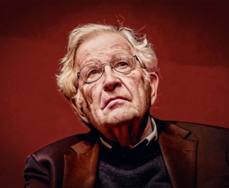 Noam Chomsky kijkt iets omhoog met een rustige, nadenkende blik, zittend tegen een effen rode achtergrond, met bril en grijs haar.