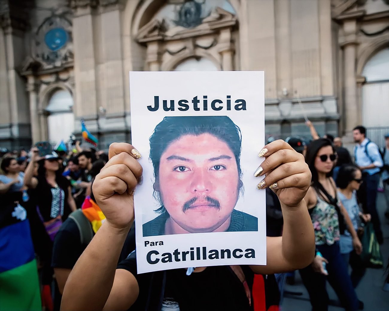 Een demonstrant houdt een bord omhoog met een geprinte portretfoto van een man en de woorden “Justicia” en “Para Catrillanca”, omringd door andere betogers.