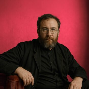 Portret van man in donkere kleding, zittend in een sobere ruimte met serieuze blik