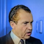 Close-up van Richard Nixon die spreekt tijdens een officiële gelegenheid, met een microfoon en blauwe achtergrond.