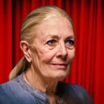 Portret van een oudere vrouw met blond haar in een lage staart, staand voor een rood gordijn.