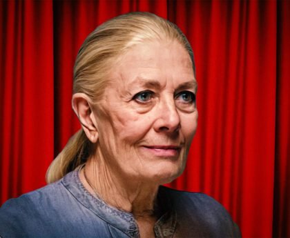 Portret van een oudere vrouw met blond haar in een lage staart, staand voor een rood gordijn.