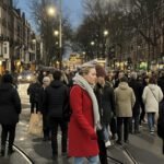 Drukke Amsterdamse straat bij schemering met voetgangers die elkaar passeren tussen tramlijnen en winkels.