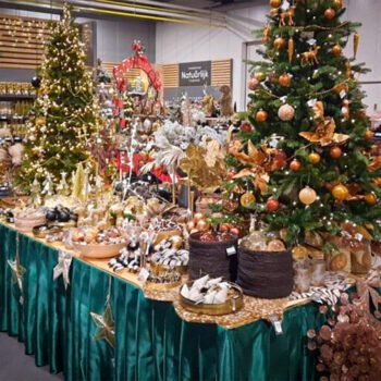 Rijk gedecoreerde kerstbomen en feesttafels vol kerstdecoratie in een winkel, met nadruk op overvloed, goudkleurige ornamenten en koopwaar.