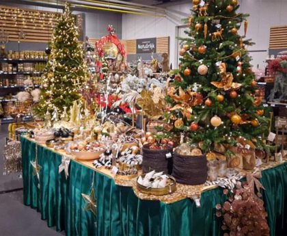 Rijk gedecoreerde kerstbomen en feesttafels vol kerstdecoratie in een winkel, met nadruk op overvloed, goudkleurige ornamenten en koopwaar.