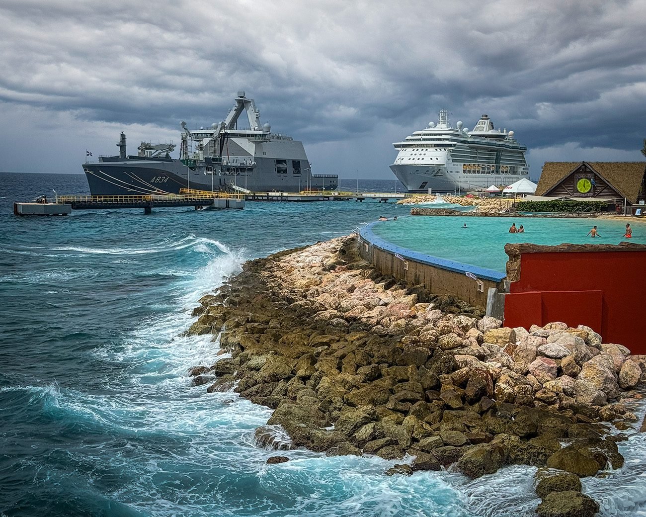 Militair schip en cruiseschip aangemeerd bij Willemstad, Curacao, terwijl donkere wolken zich samenpakken boven zee.