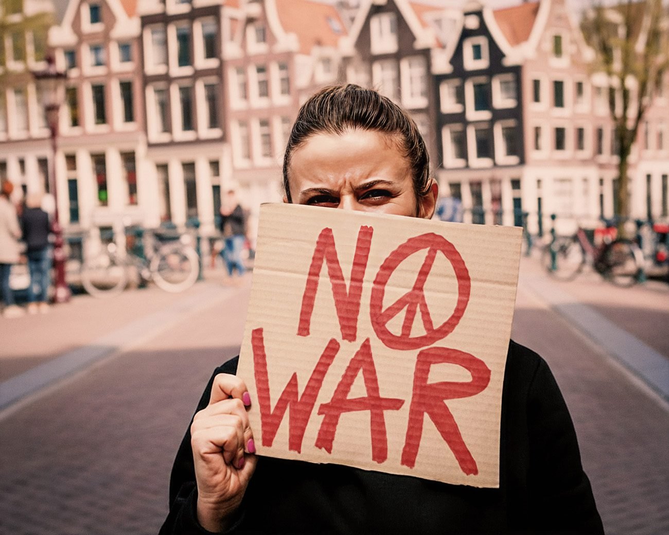 Persoon houdt een kartonnen bord met de tekst “No War” omhoog in een Amsterdamse straat