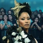 Nicki Minaj op een podium kijkt strak vooruit terwijl een zichtbaar emotioneel publiek op de achtergrond toekijkt.
