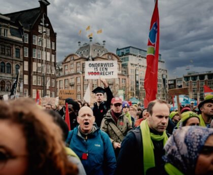 Studenten en docenten op de Dam tijdens een protest tegen bezuinigingen op het hoger onderwijs, met spandoeken en uitzicht op het plein.