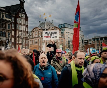 Studenten en docenten op de Dam tijdens een protest tegen bezuinigingen op het hoger onderwijs, met spandoeken en uitzicht op het plein.
